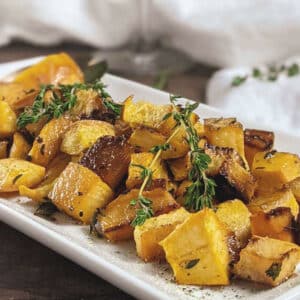 A white rectangular plate with roasted butternut squash, potatoes, and roasted rutabaga, all garnished with fresh herbs.