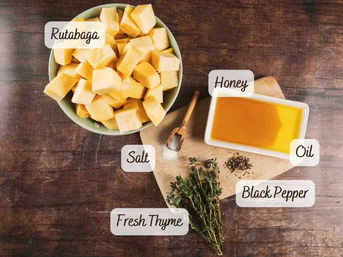 Cubed roasted rutabaga in a bowl sits beside a cutting board with fresh thyme, salt, black pepper, honey, and oil on a wooden surface.