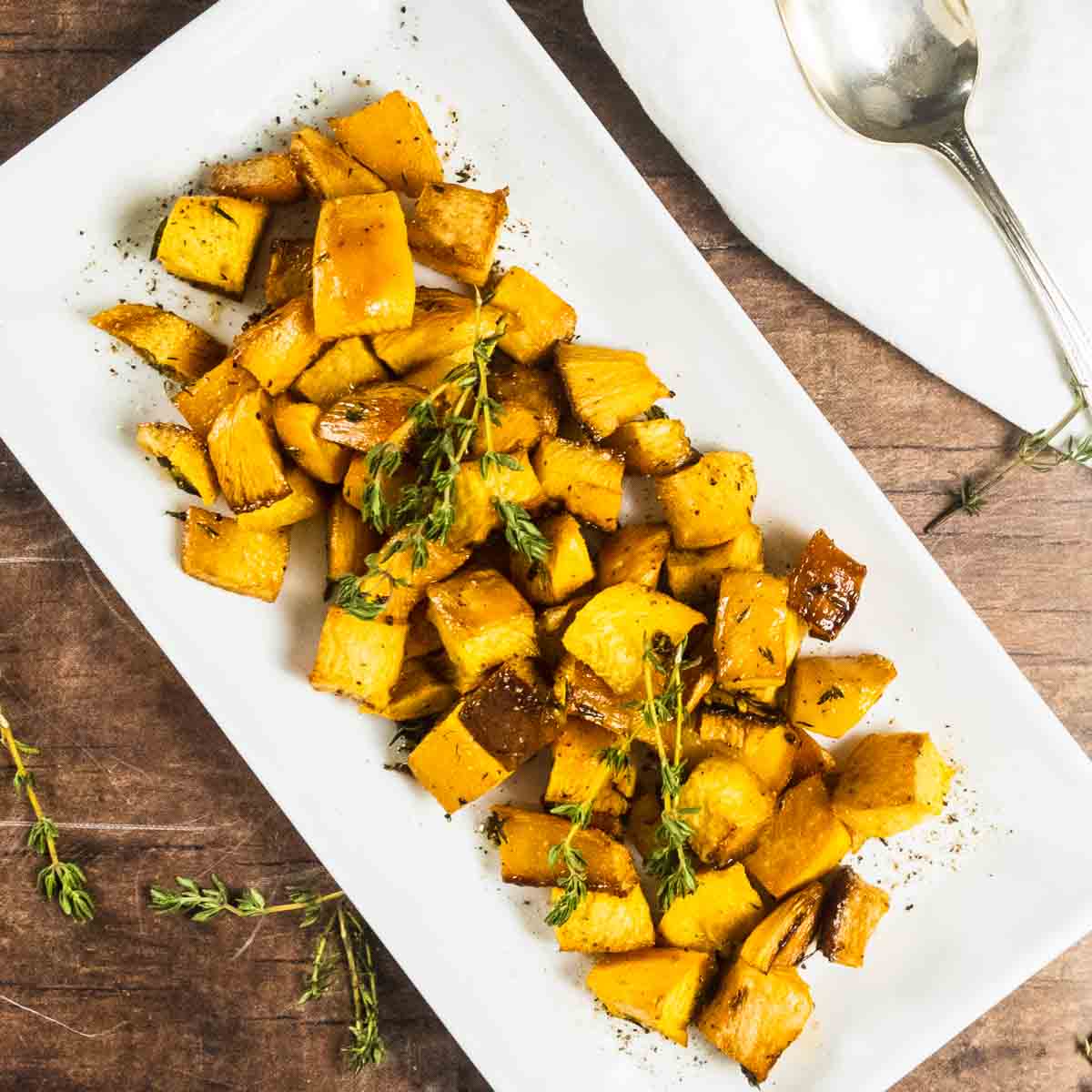 A white rectangular plate with roasted rutabaga squash cubes, seasoned to perfection and garnished with sprigs of thyme, sits next to a silver spoon.