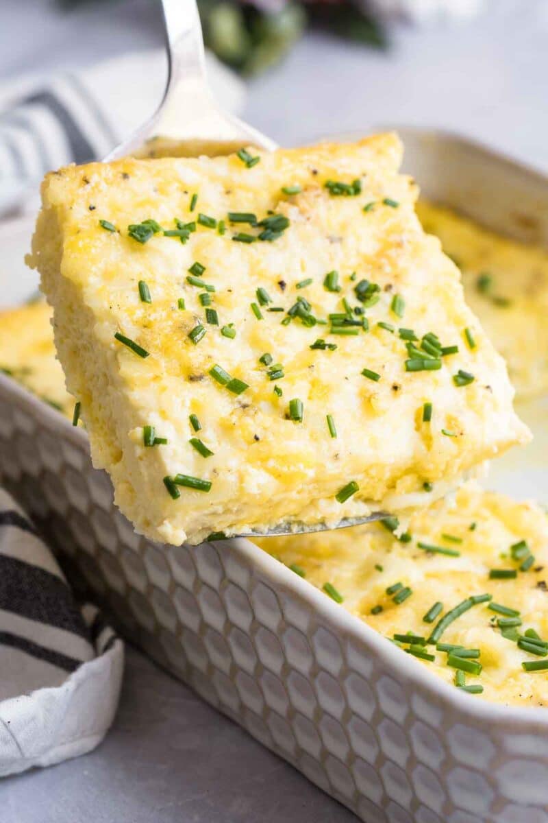 A slice of gluten free egg breakfast casserole topped with chopped chives is being lifted from a baking dish.
