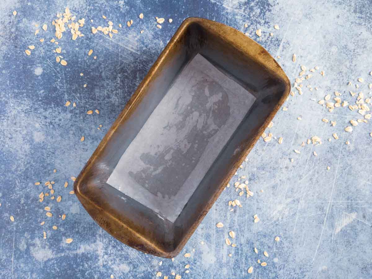 A rectangular metal loaf pan sits on a blue textured surface with scattered oats around it.