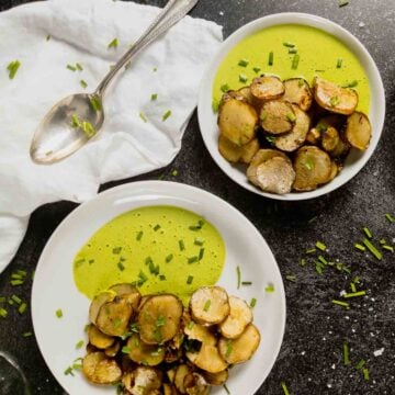 Two bowls of roasted and sauteed sunchokes topped with chopped chives, served with a vibrant green sauce, sit beside a spoon and a white napkin on a dark surface.