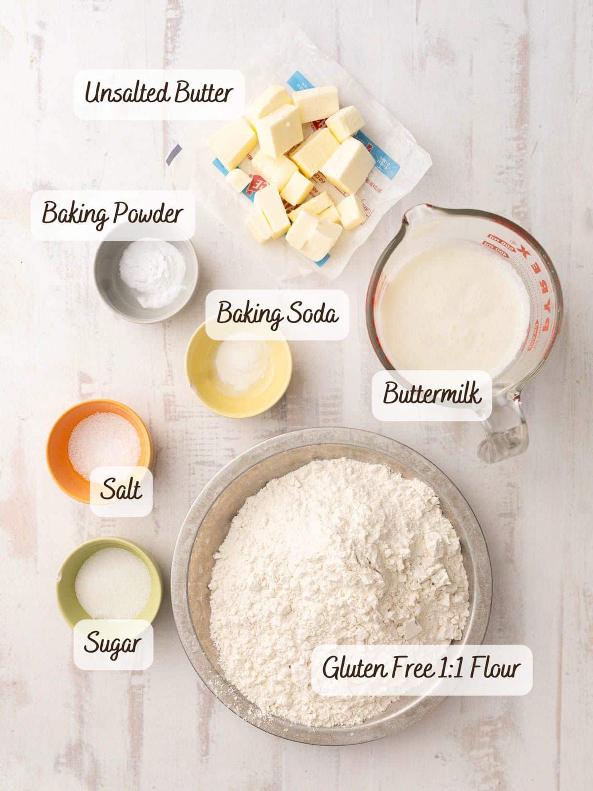 Overhead view of gluten-free soda bread ingredients: cubed unsalted butter, baking powder, baking soda, buttermilk, salt, sugar, and 1:1 gluten-free flour in bowls and a measuring cup on a light surface.