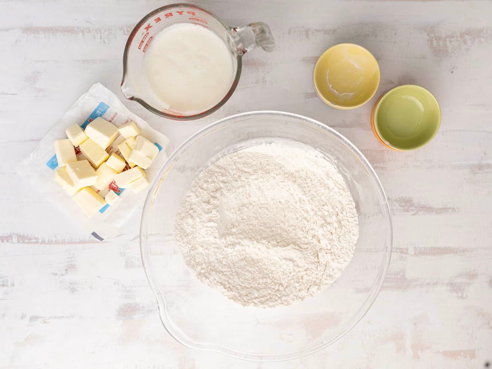 Ingredients for gluten-free soda bread on a table: a bowl of flour, a glass container with milk, several butter pieces on a wrapper, and two small empty bowls.