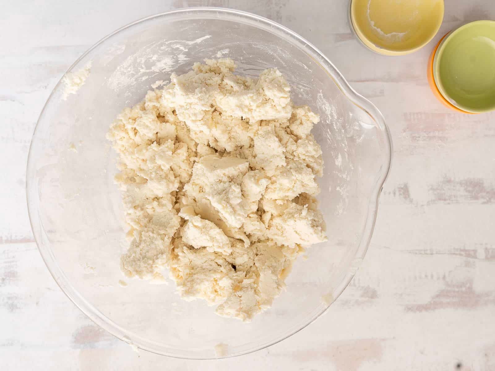 A clear bowl with gluten-free soda bread dough rests on a light surface. Two small empty bowls, one yellow and one green, are partially visible at the top right.