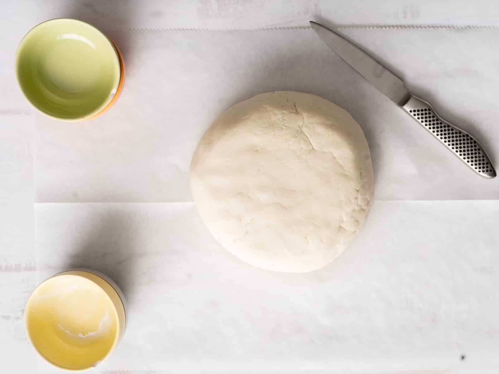 A ball of gluten-free soda bread dough rests on parchment paper, flanked by a knife and two small empty bowls.