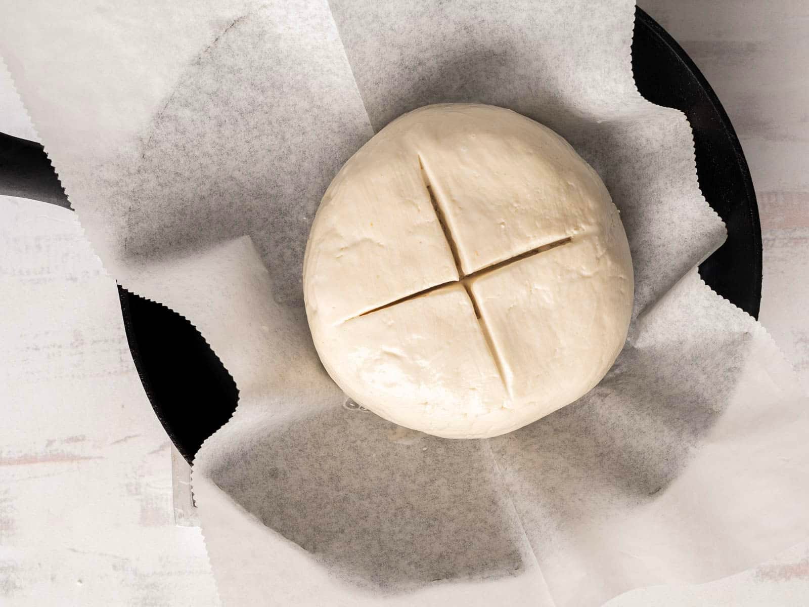 A ball of gluten-free soda bread dough with a cross cut on top rests on parchment paper in a black pan.