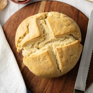 A round loaf of gluten-free soda bread sits on a wooden board, accompanied by a serrated knife and a folded cloth.
