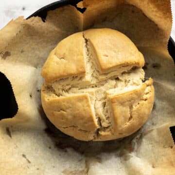 A round loaf of gluten-free soda bread with a cross cut on top, baked on parchment paper in a black skillet.