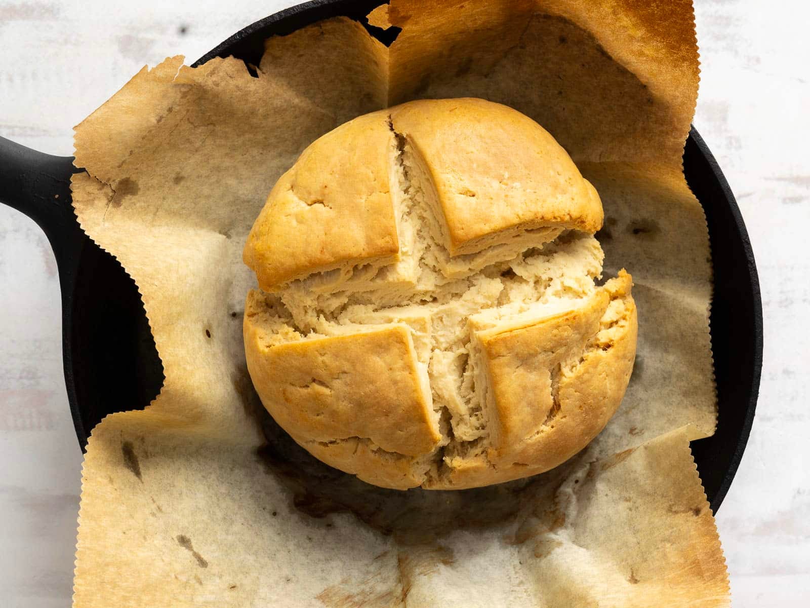A round loaf of gluten-free soda bread with a cross cut on top, baked on parchment paper in a black skillet.