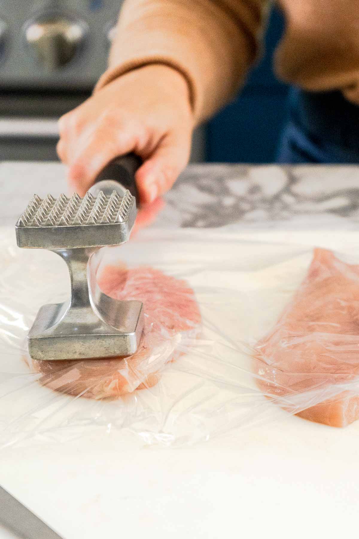 chicken breast covered in plastic wrap being flattened with meat mallet
