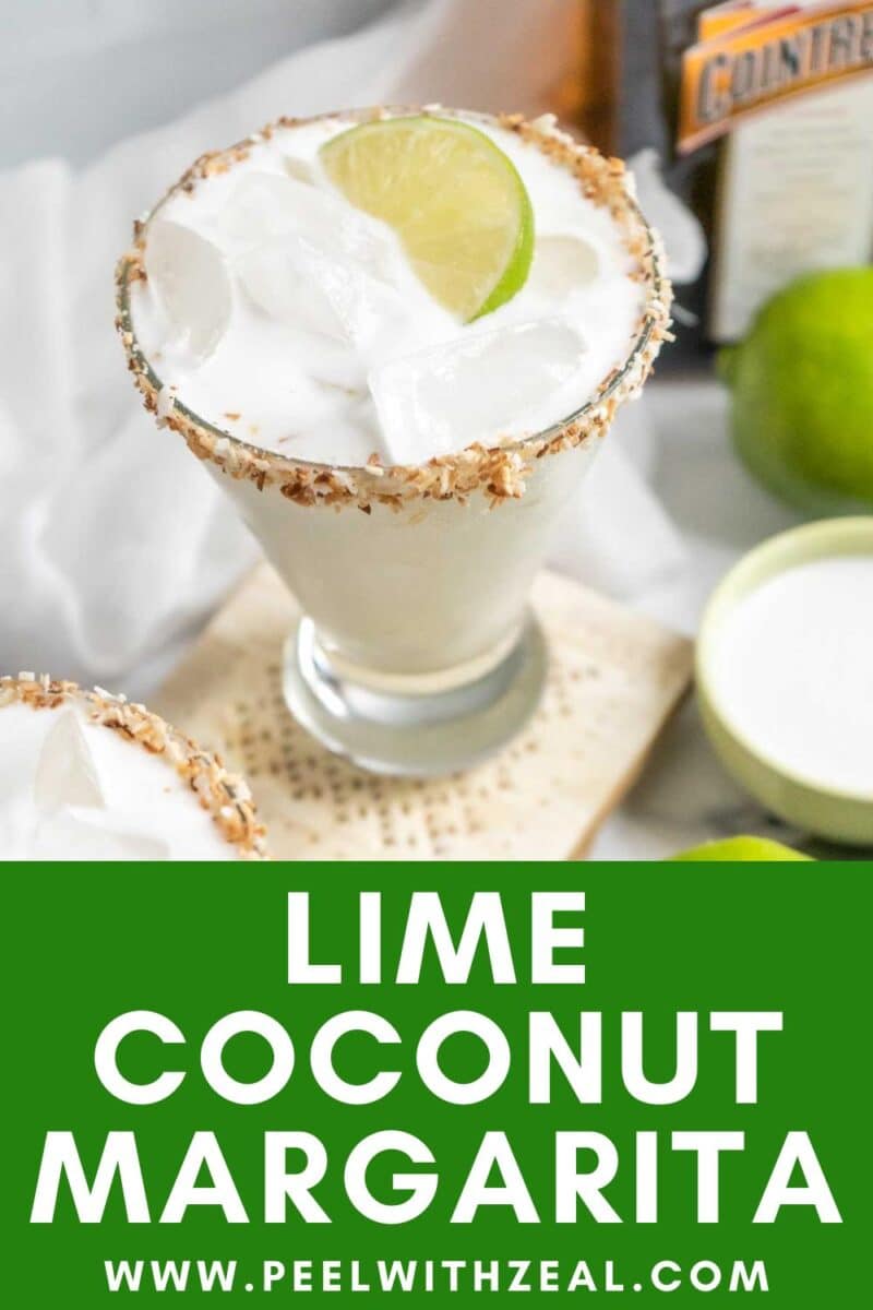 A glass of coconut margarita with ice, garnished with a lime wedge and toasted coconut rim, sits on a table surrounded by fresh limes and a bottle in the background.