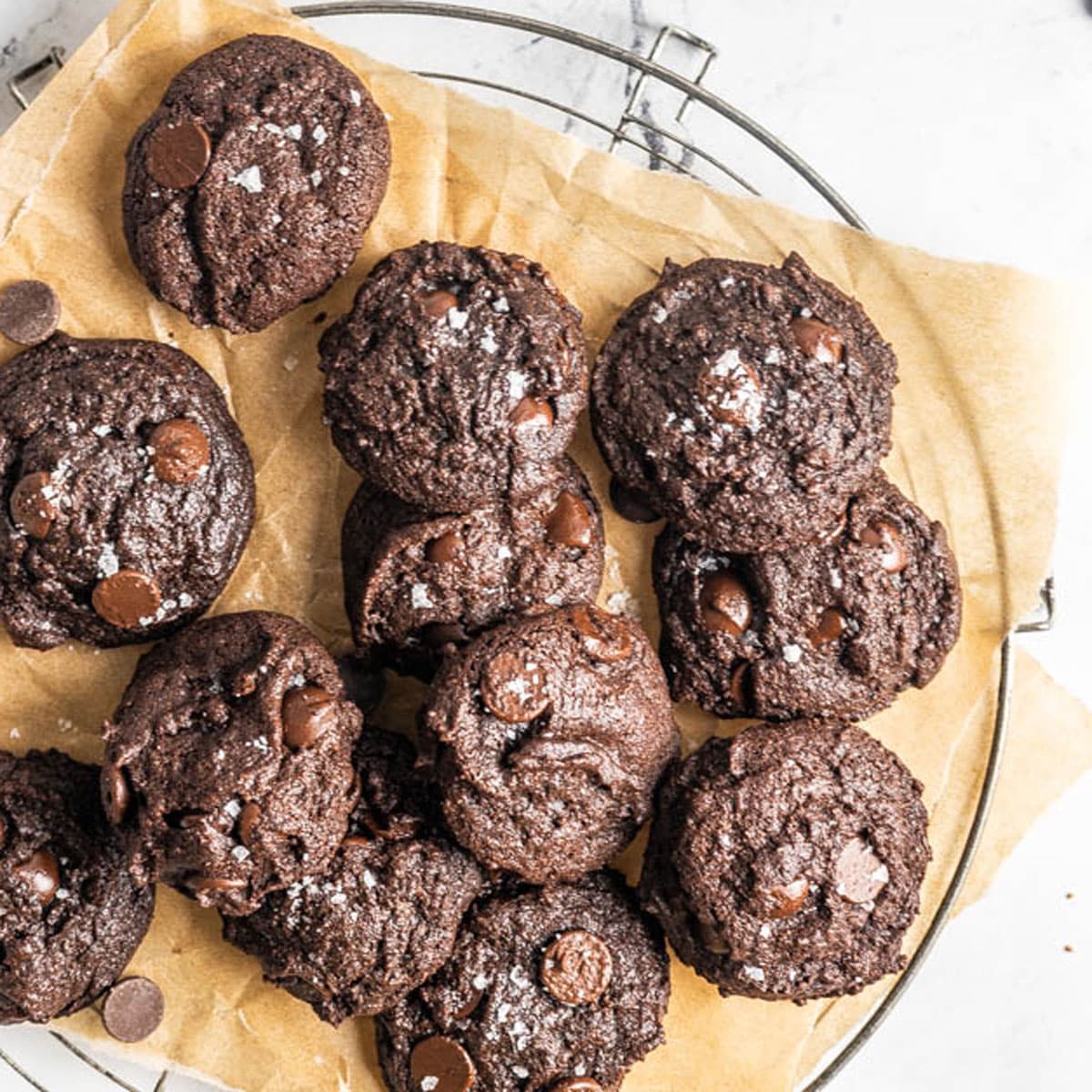 A wire rack with several chocolate cookies made from oat flour recipes, topped with chocolate chips and a sprinkle of flaky sea salt, placed on parchment paper.