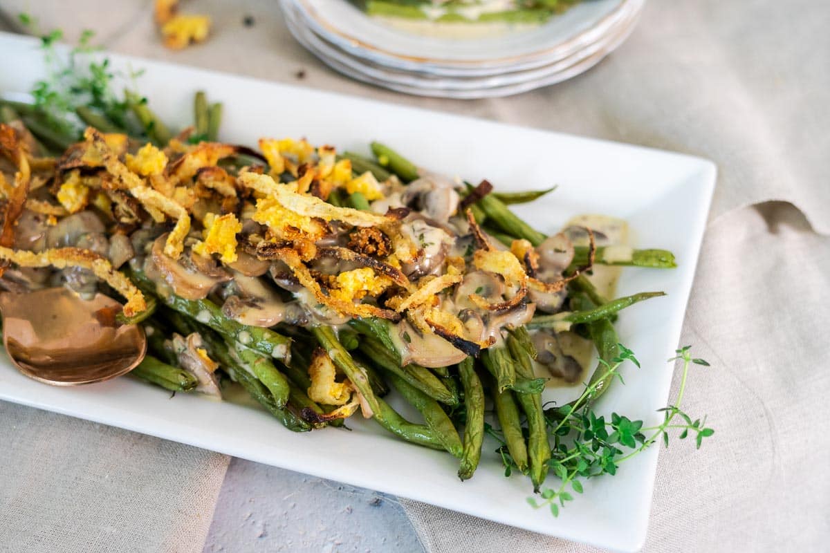 A rectangular white plate with roasted green beans, sautéed mushrooms, crispy fried onions, and a creamy sauce, garnished with fresh herbs-a modern green bean casserole reimagined for today's table.