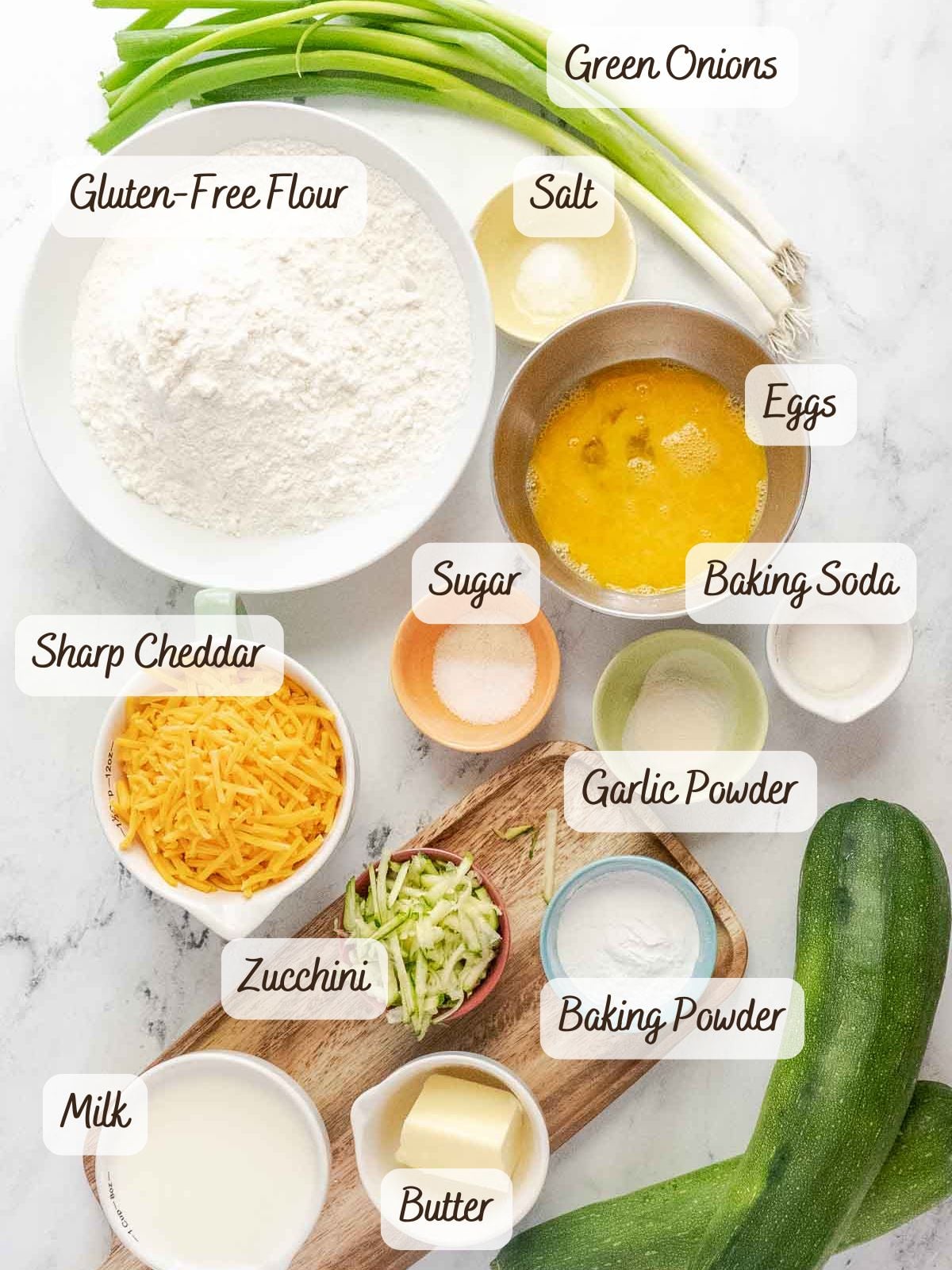 Ingredients for gluten free zucchini bread are arranged on a white surface, including gluten-free flour, green onions, eggs, cheddar, zucchini, milk, butter, and various seasonings.