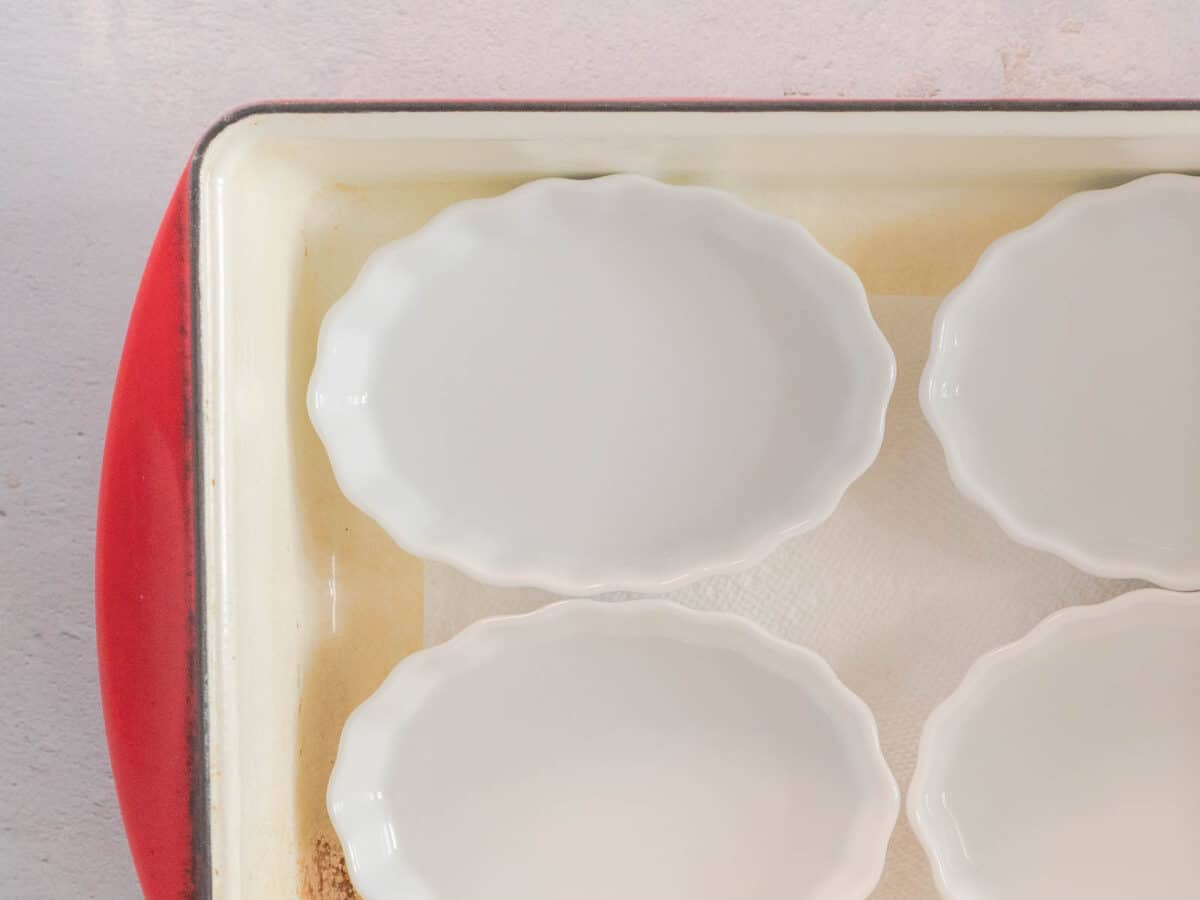 A top-down view of four empty white ramekins in a red-bordered baking dish lined with paper towels, ready to be filled for maple creme brulee.
