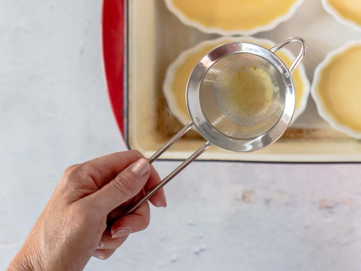 A hand holds a fine mesh strainer above ramekins filled with pale yellow liquid, likely preparing maple creme brulee for baking.