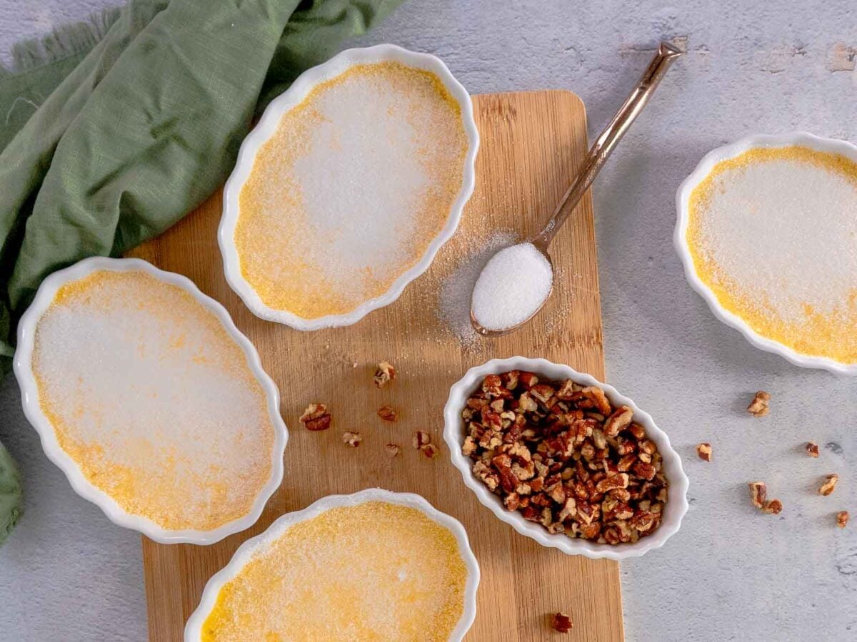 Four ramekins of maple creme brulee with a golden sugar crust sit on a wooden board, alongside a spoon with sugar, a bowl of chopped pecans, and a green cloth nearby.