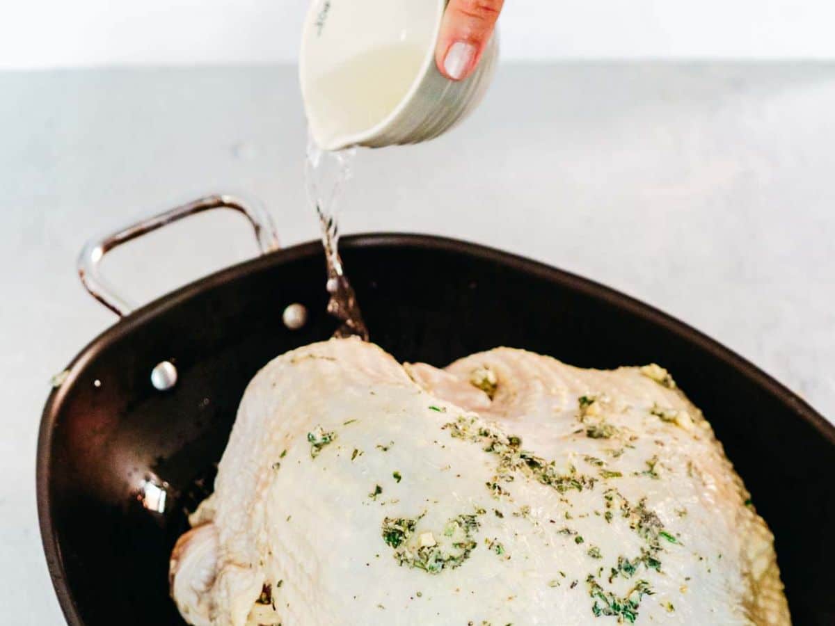 A hand pours liquid from a small bowl over a seasoned bone in turkey breast nestled in a black roasting pan.