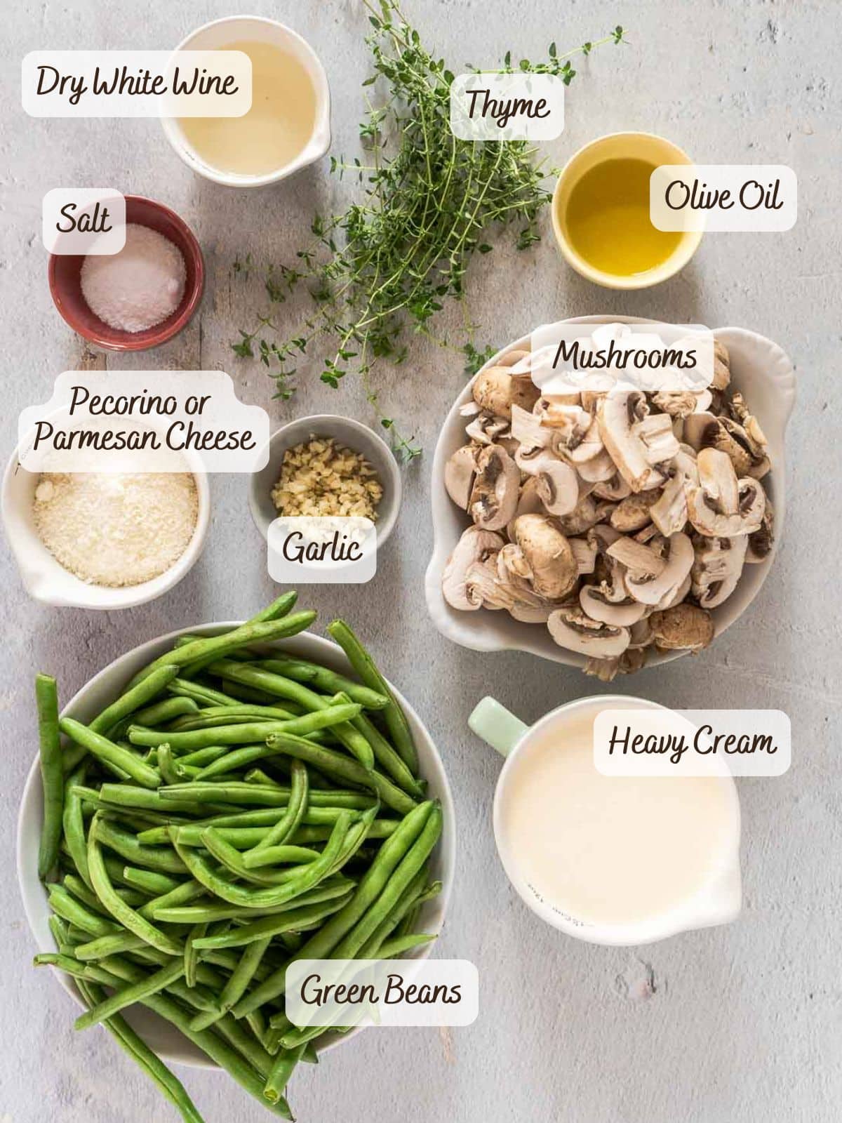 Assorted ingredients on a table for a modern green bean casserole, including green beans, mushrooms, garlic, salt, dry white wine, thyme, olive oil, heavy cream, and grated cheese.