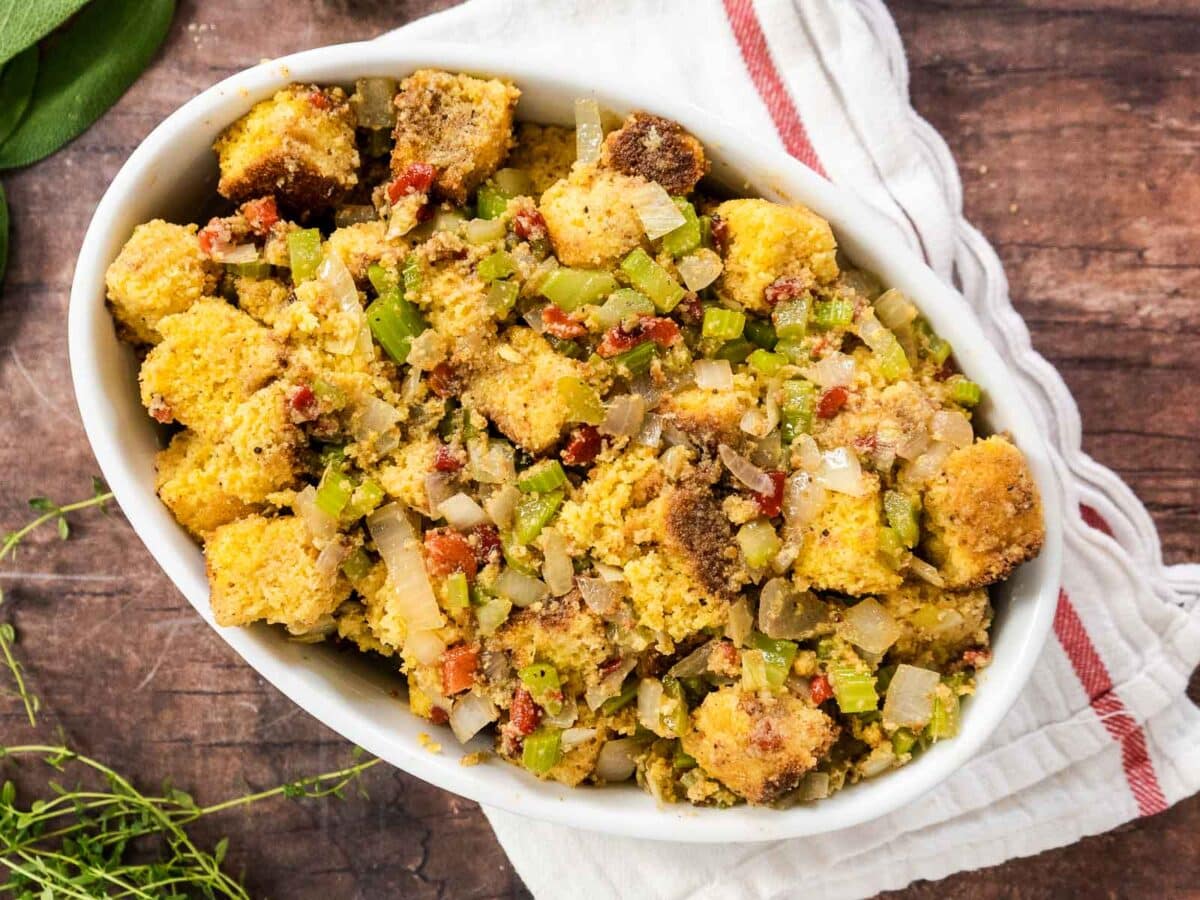 Oval dish filled with gluten-free cornbread stuffing containing diced celery, onions, and small pieces of bacon, placed on a wooden surface with a white cloth underneath.