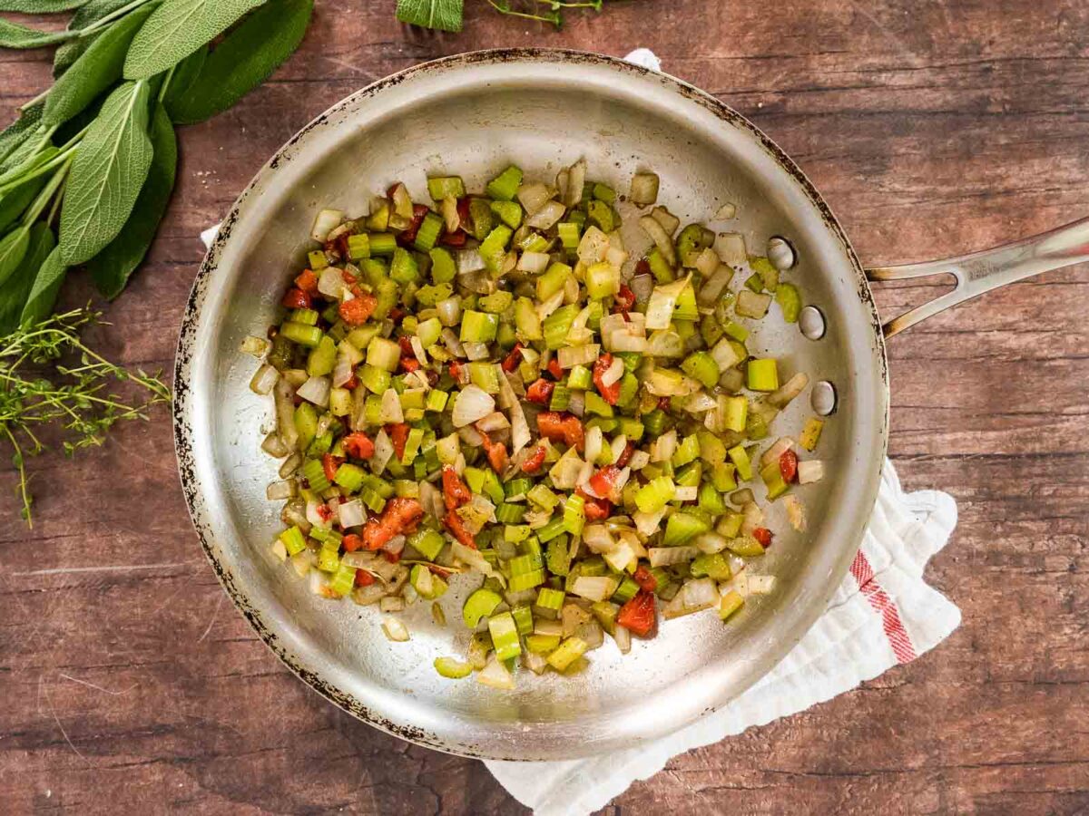 A stainless steel skillet on a wooden surface holds sautéed diced celery, onions, and red bell peppers-delicious ingredients for gluten-free cornbread stuffing. Fresh herbs and a white towel are nearby.