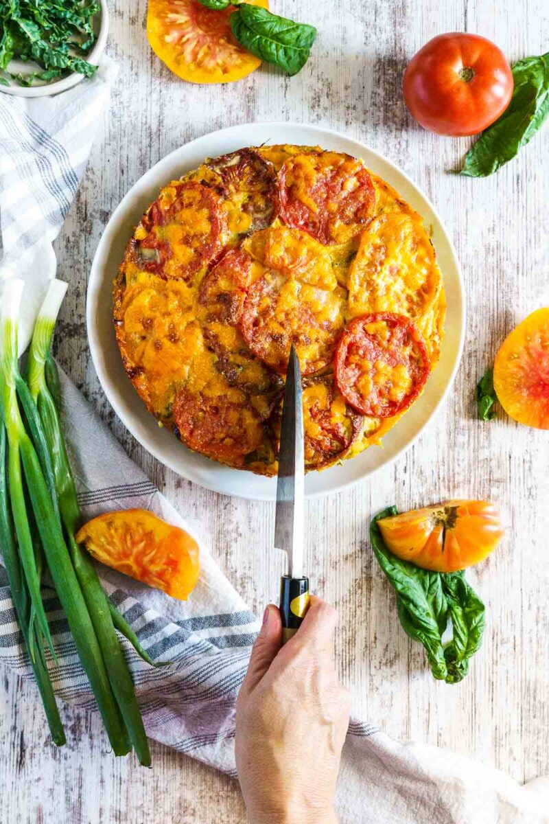 A hand cutting a slice of a baked tomato and cheese pie on a white plate, surrounded by fresh tomatoes, green onions, and basil on a rustic wooden table.