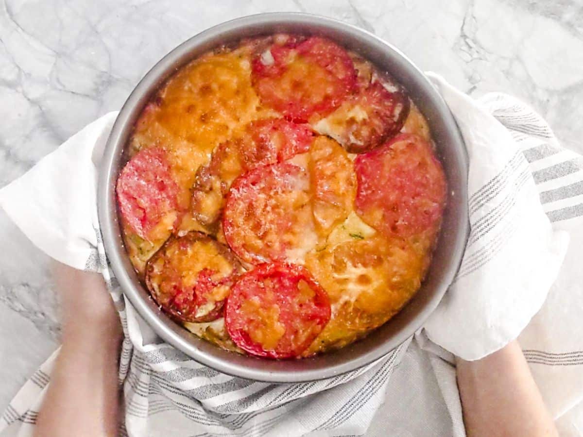 A crustless tomato pie-this round baked dish is topped with sliced tomatoes and melted cheese, held with a towel over a marble surface.