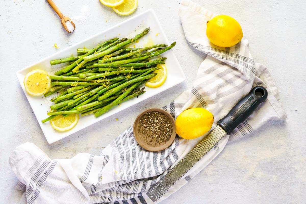 Plate of sous vide asparagus with lemon wedges on a striped cloth. Nearby are a zest grater, whole lemons, a wooden spoon, and a small bowl of seasoning.