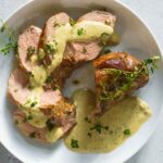 Sliced sous vide pork with creamy herb sauce and fresh chopped herbs served on a white plate.