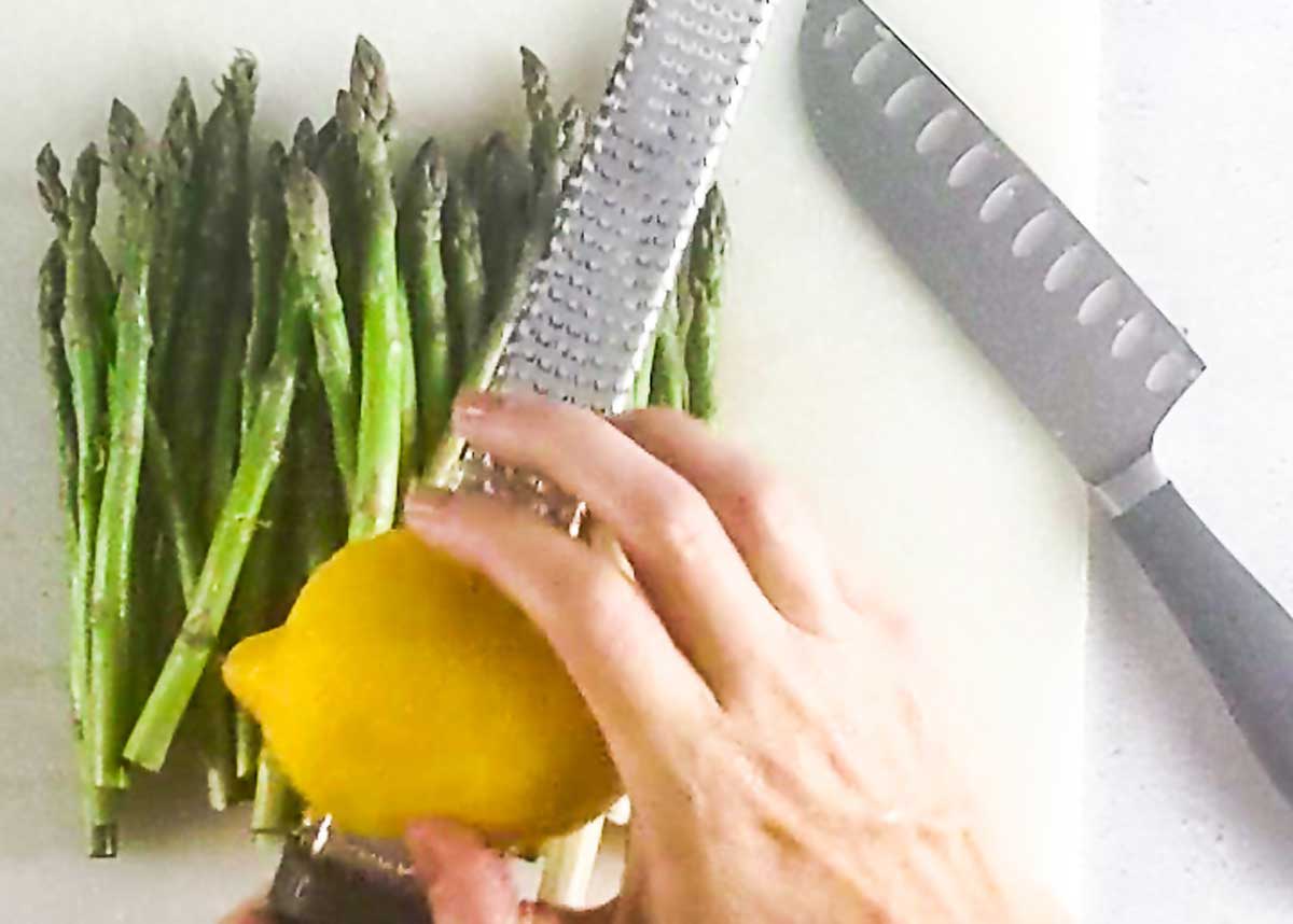 Hands grating a lemon zest onto asparagus on a cutting board next to a knife.