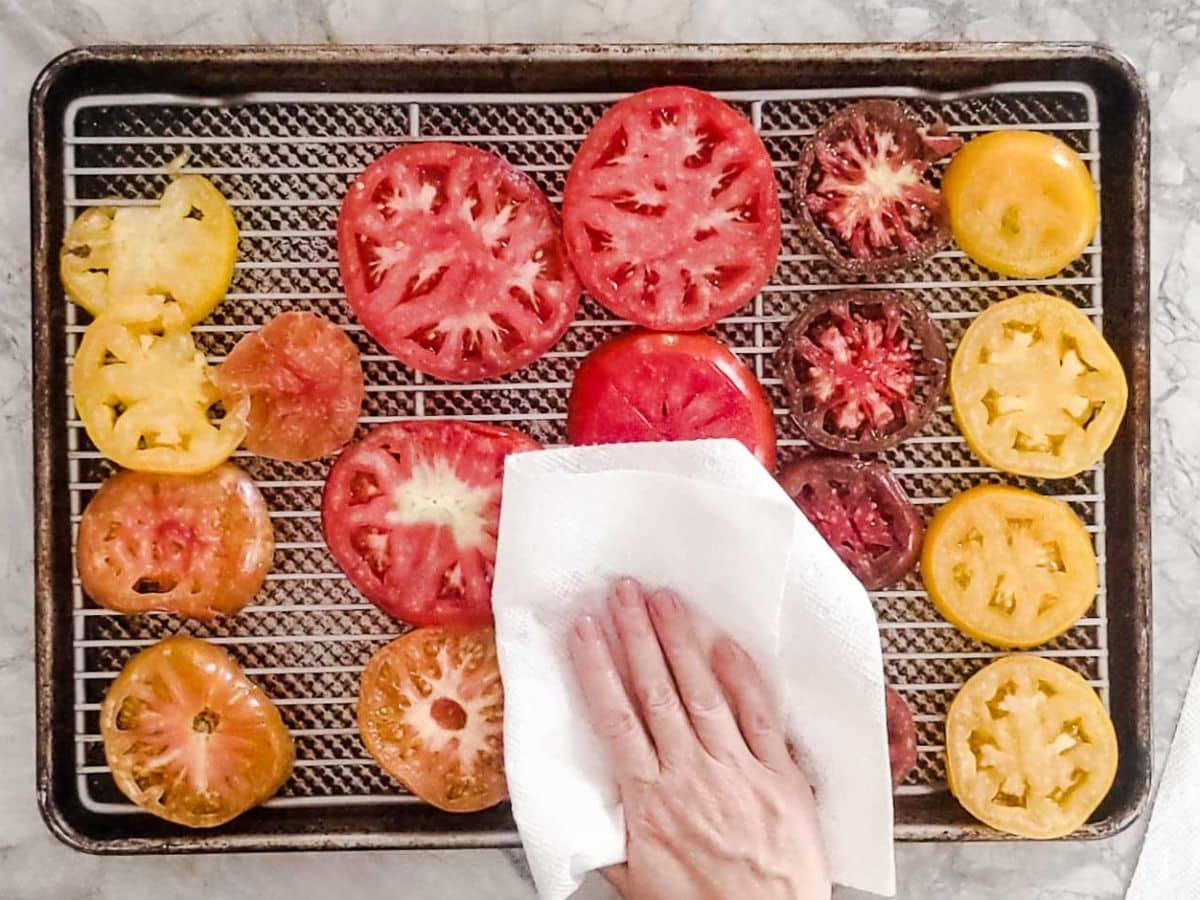 A hand patting sliced tomatoes with a paper towel on a wire rack set over a baking sheet, preparing them for a delicious crustless tomato pie.