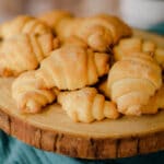 A wooden platter filled with several golden-brown, flaky gluten free crescent rolls placed on a teal cloth.
