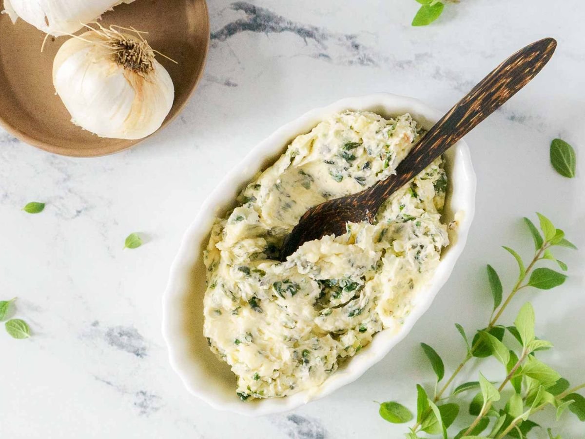 Oval dish filled with herb butter, perfect for spreading on gluten free garlic bread, with a wooden spoon inside. Fresh herbs and garlic bulbs are on the marble surface nearby.