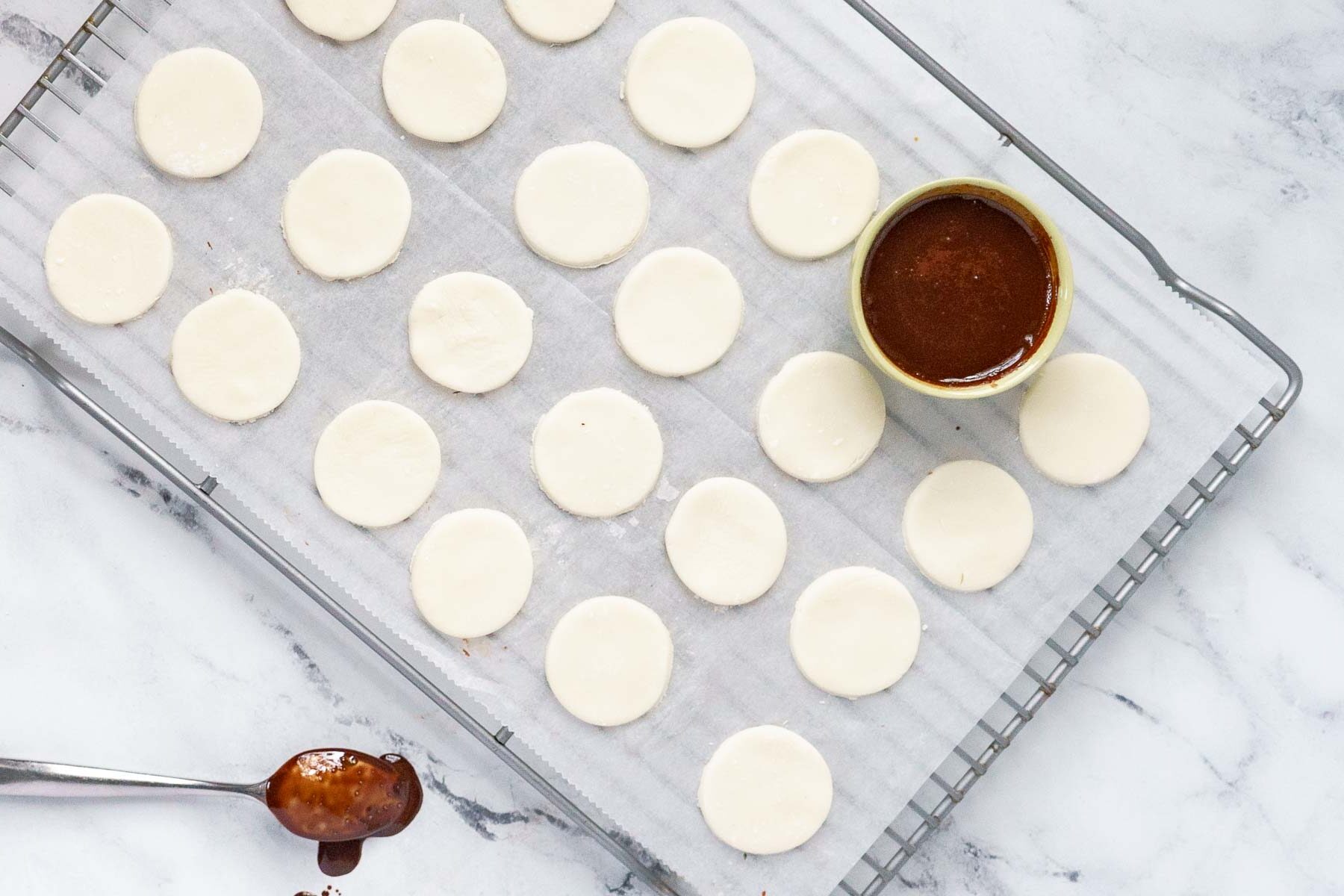 Round pastry circles arranged on parchment paper on a cooling rack, with a bowl of chocolate filling and a spoon with chocolate nearby.