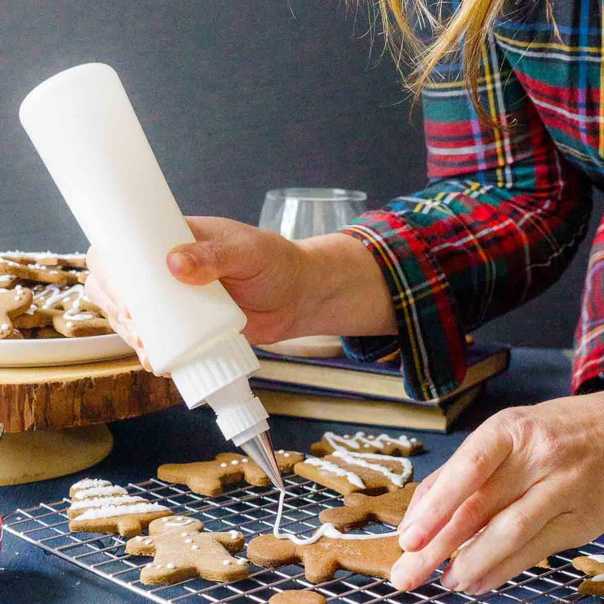 A person decorates gingerbread cookies with royal icing made from meringue powder using a squeeze bottle; cookies rest on a wire rack and a plate.
