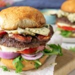 A wagyu beef hamburger with lettuce, tomato, red onion, bacon, blue cheese, and a beef patty on a bun sits on parchment paper; a blackened chicken sandwich is blurred in the background.
