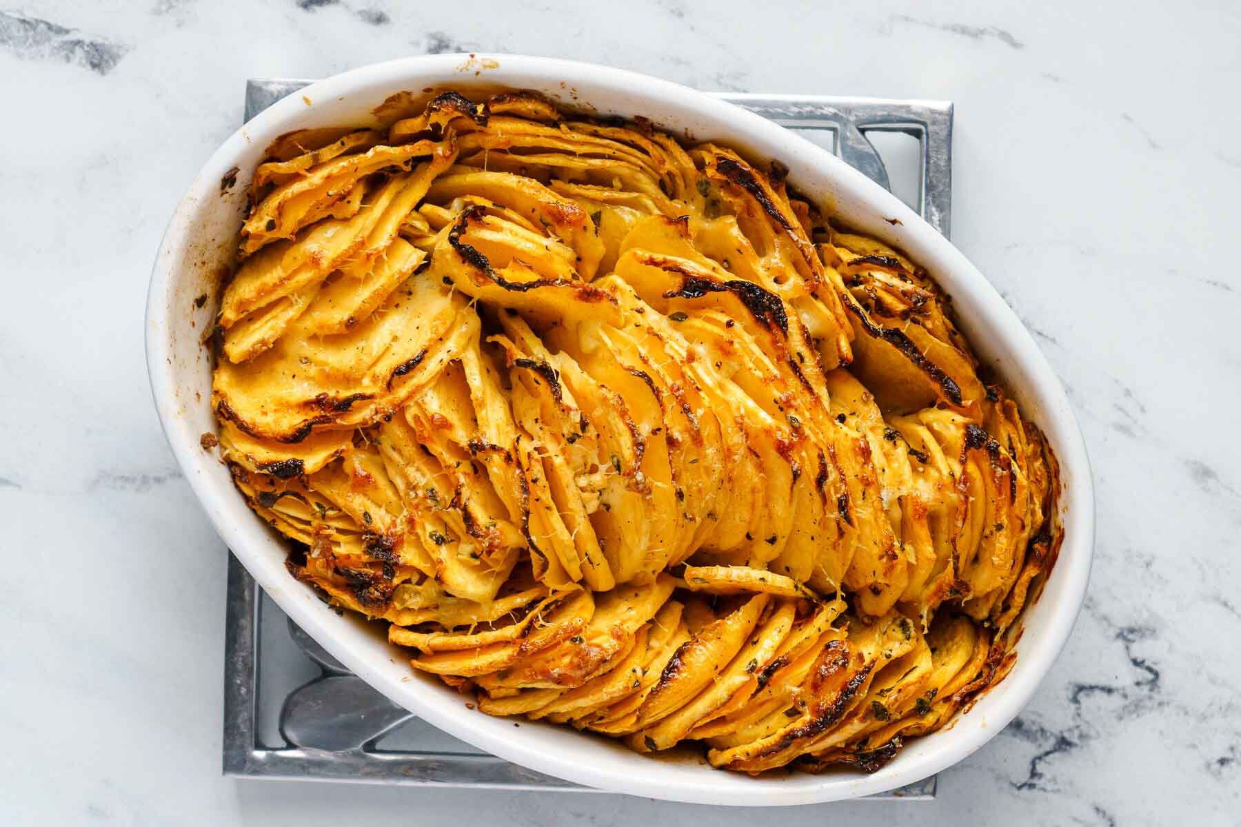 An oval baking dish filled with thinly sliced, golden-brown roasted potatoes and baked rutabaga, arranged vertically in overlapping layers.