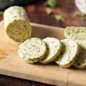A log of Herb and Garlic Compound Butter is partially sliced on a wooden cutting board, with several round slices lying in front of it.