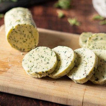 A log of Herb and Garlic Compound Butter is partially sliced on a wooden cutting board, with several round slices lying in front of it.