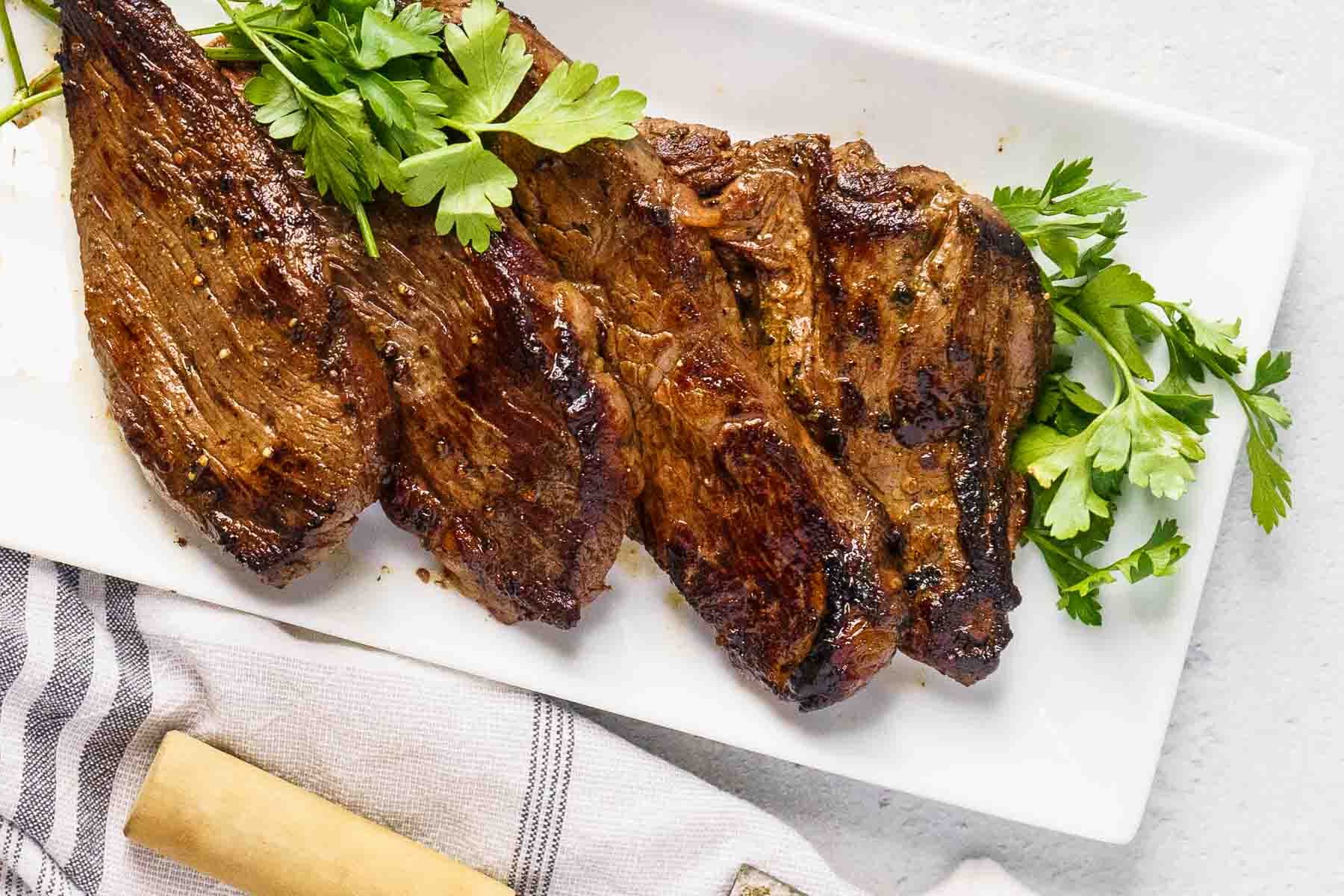 Three grilled bavette steaks garnished with fresh parsley on a white rectangular plate, placed next to a striped napkin.