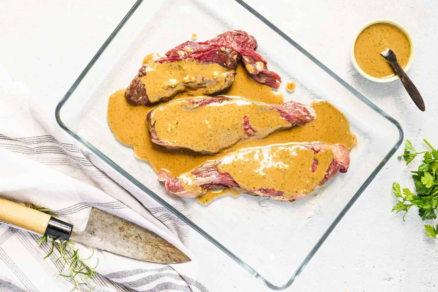 Three raw bavette steaks in a glass dish are covered with a French marinade; a bowl of sauce, knife, herbs, and striped towel are nearby.