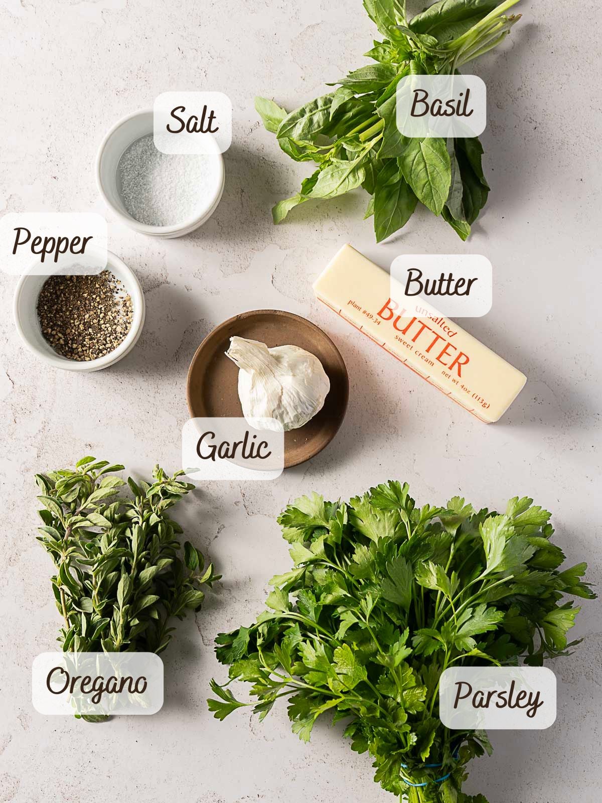 A countertop displays labeled ingredients for Herb and Garlic Compound Butter: basil, parsley, oregano, garlic, butter, salt, and pepper.