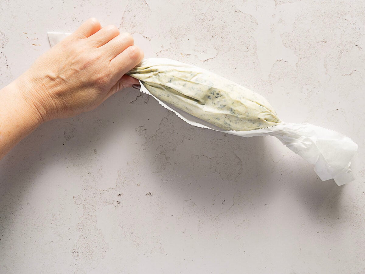A hand twists the ends of a sheet of parchment paper enclosing a log of herb and garlic compound butter on a light textured surface.