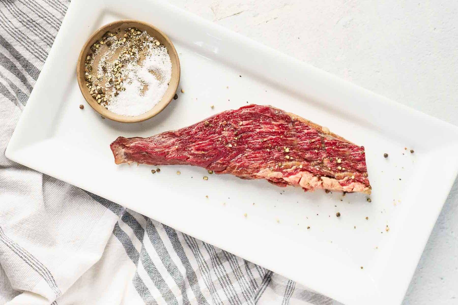 A raw cut of bavette steak with visible marbling is placed on a rectangular white plate next to a small dish of salt and pepper.
