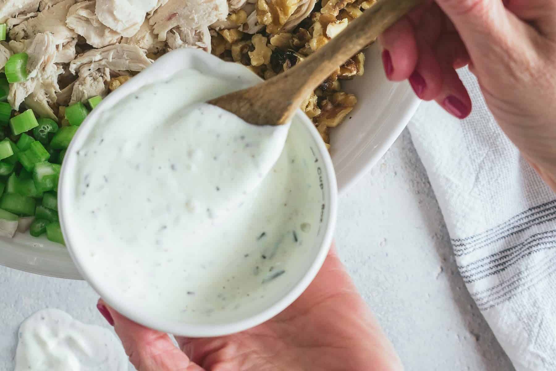 A person mixes creamy herb dressing with a wooden spoon for the cranberry chicken salad. Nearby are chopped green peppers and shredded chicken in a bowl. A small amount of dressing has spilled onto the counter.