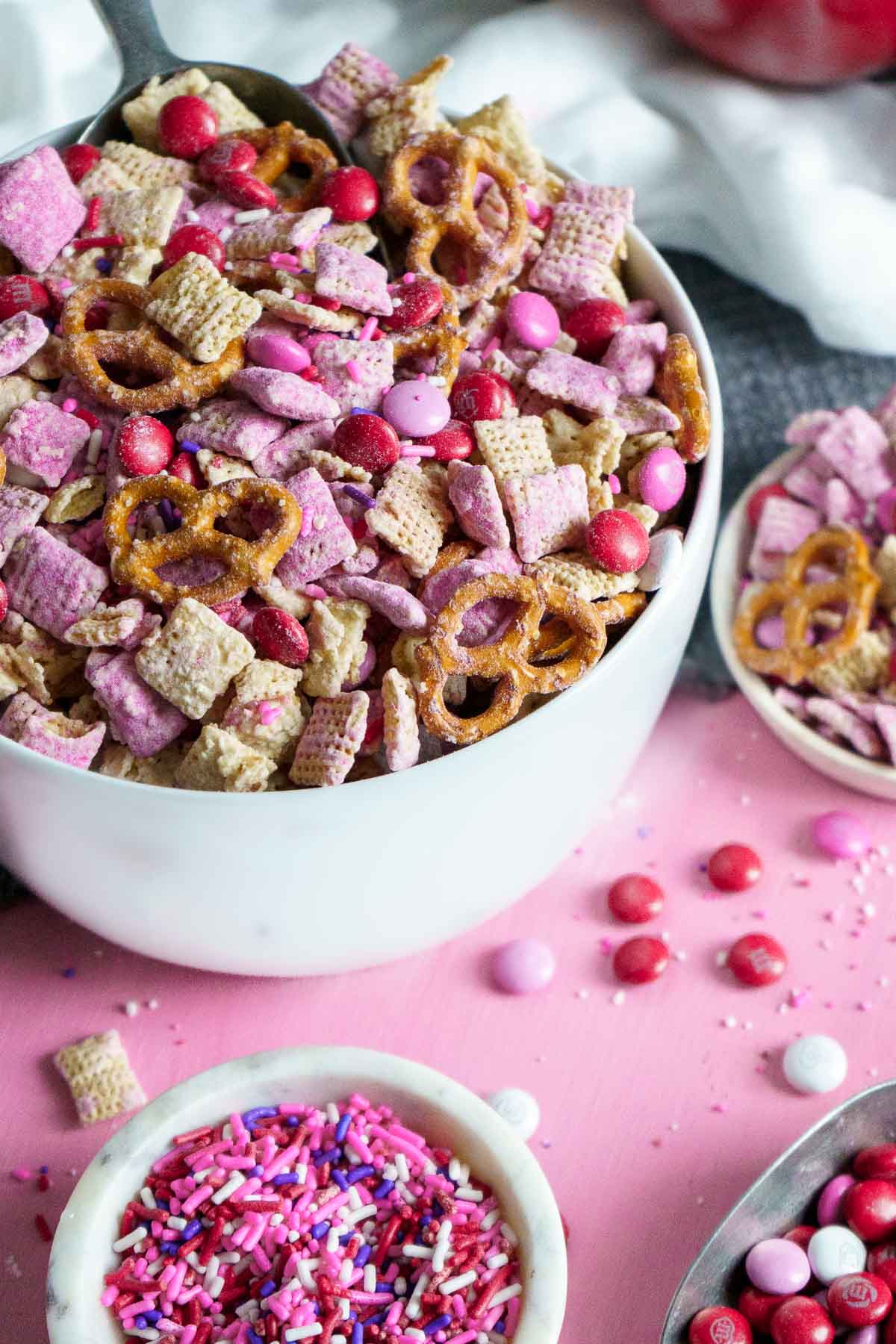 bowl of pink puppy chow mix surrounded by sprinkles and M&M candies.
