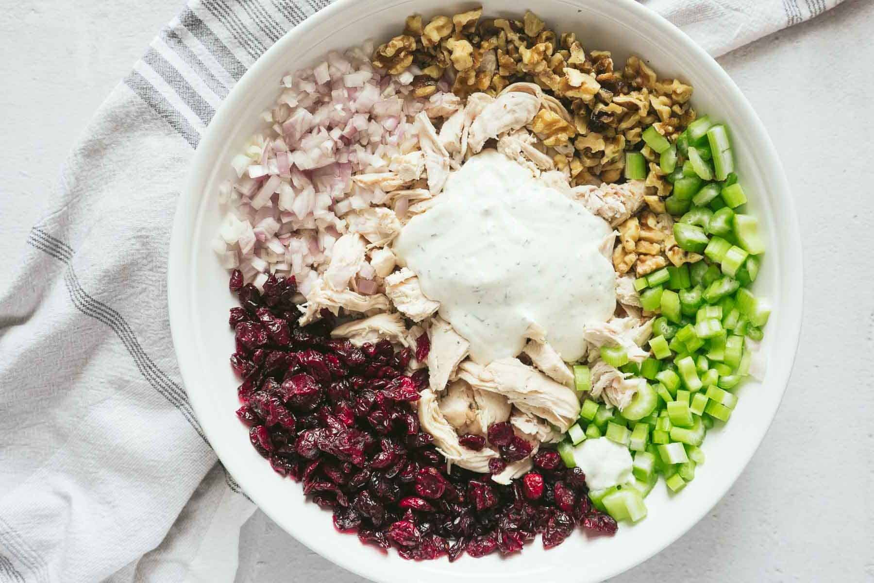 A vibrant cranberry chicken salad bowl features chopped chicken, crunchy walnuts, crisp celery, tangy cranberries, and sliced shallots, all topped with a dollop of dressing and set on a gray-striped cloth.