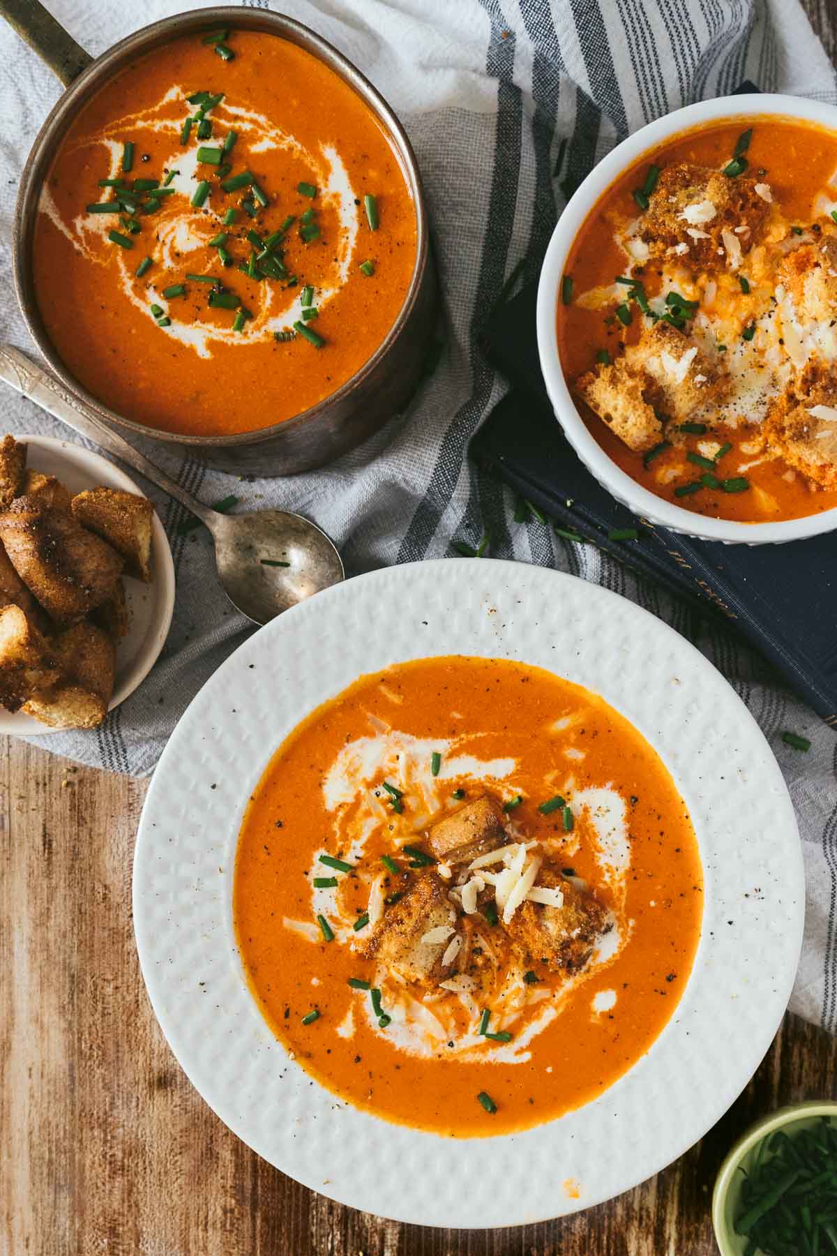 three bowls of red pepper soup topped with gouda and drizzled with cream and chives.