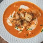 A bowl of roasted red pepper soup garnished with cream, croutons, shredded gouda cheese, chopped chives, and black pepper,